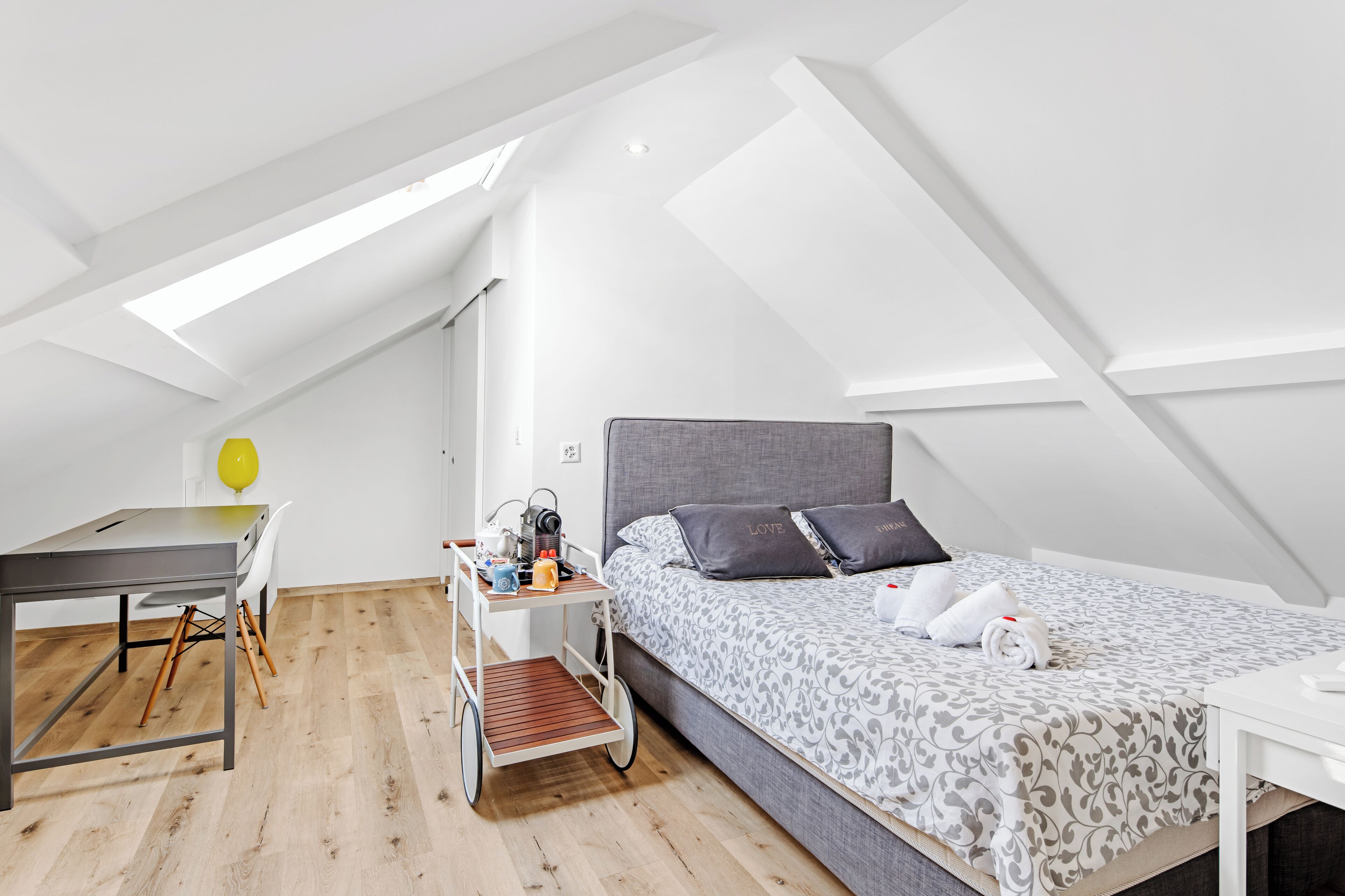A modern, compact room with a loft-style ceiling, a bed with patterned bedding, a desk with a chair, and a small cart with items.