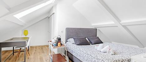 A modern, compact room with a loft-style ceiling, a bed with patterned bedding, a desk with a chair, and a small cart with items.