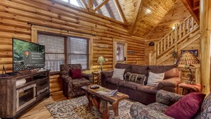 A log cabin interior with a wooden ceiling, a television displaying a nature scene, a fireplace, and a cozy seating area with patterned cushions.