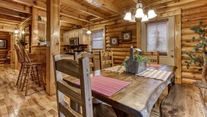 A log cabin interior with a wooden dining table, chairs, a kitchen area, and a ceiling with exposed beams.