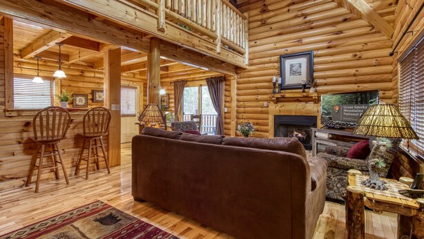 A log cabin interior with a wooden staircase, a fireplace, a sofa, and a dining area.