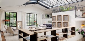 Ein modernes Wohnzimmer mit einer großen Holzbank, einer Couch und einem Esstisch. Große Fenster mit Vorhängen, eine Treppe und gerahmte Kunstwerke an den Wänden.
