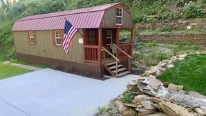 A small, single-story cabin with a porch and a flag, surrounded by a stone wall and greenery.