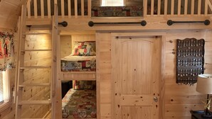 A cozy cabin interior with a loft bedroom, wooden floors, a red rug, and a wall-mounted lamp.