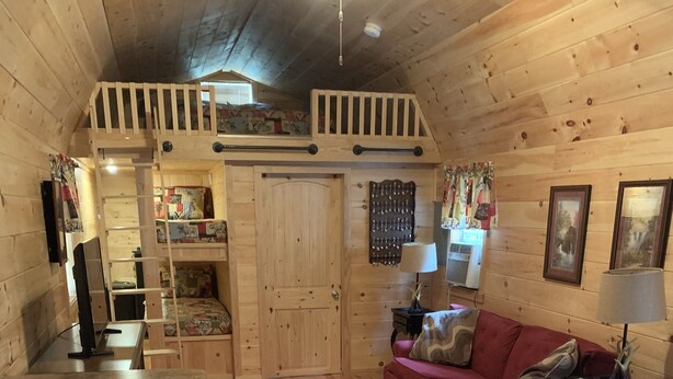 A cozy cabin interior with a wooden interior, a red couch, a small kitchen area, and a loft bedroom.