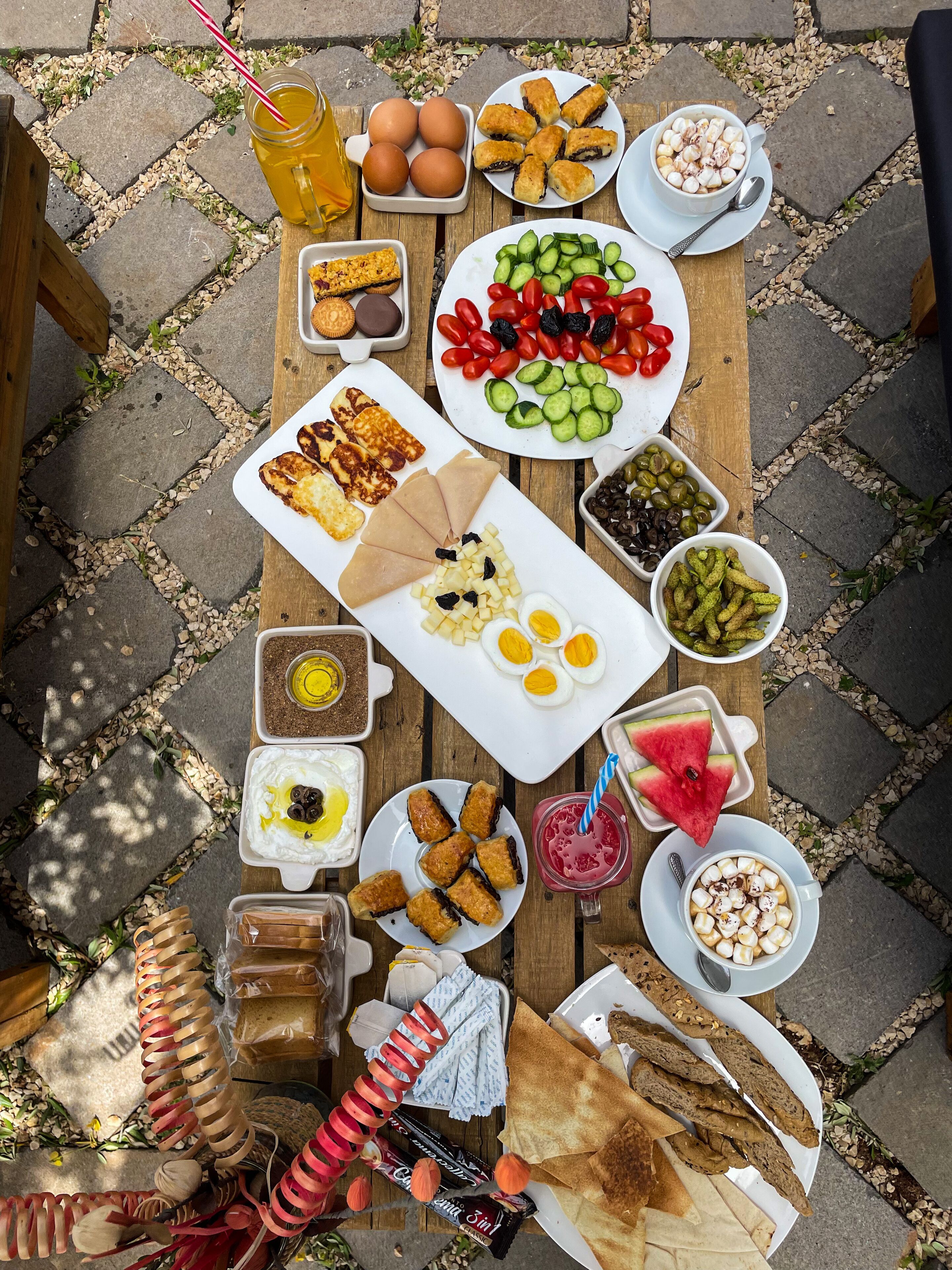 A variety of food items and drinks arranged on a wooden table outdoors.
