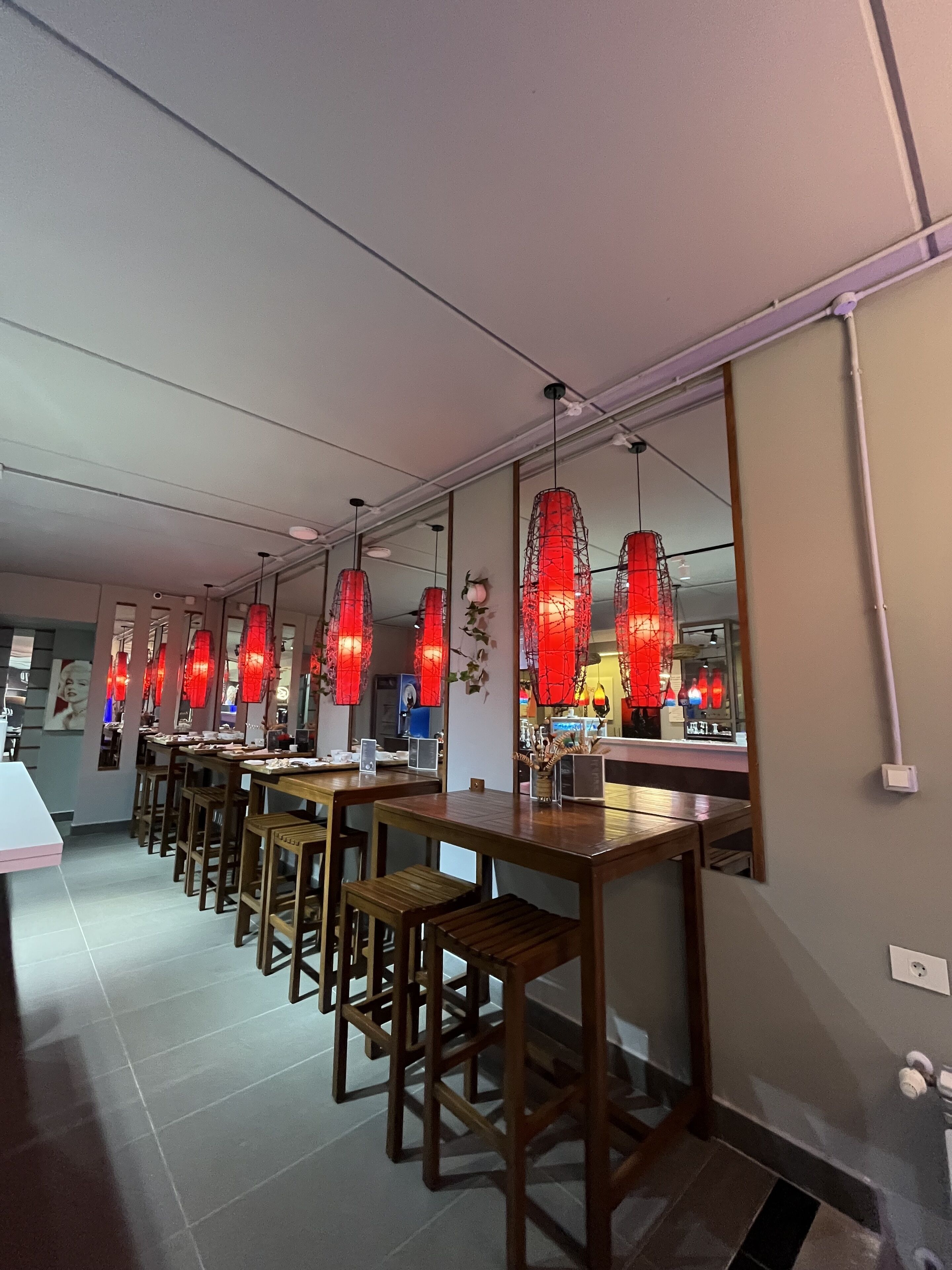 Un restaurante con mesas y sillas de madera, luces colgantes rojas y suelo de baldosas.