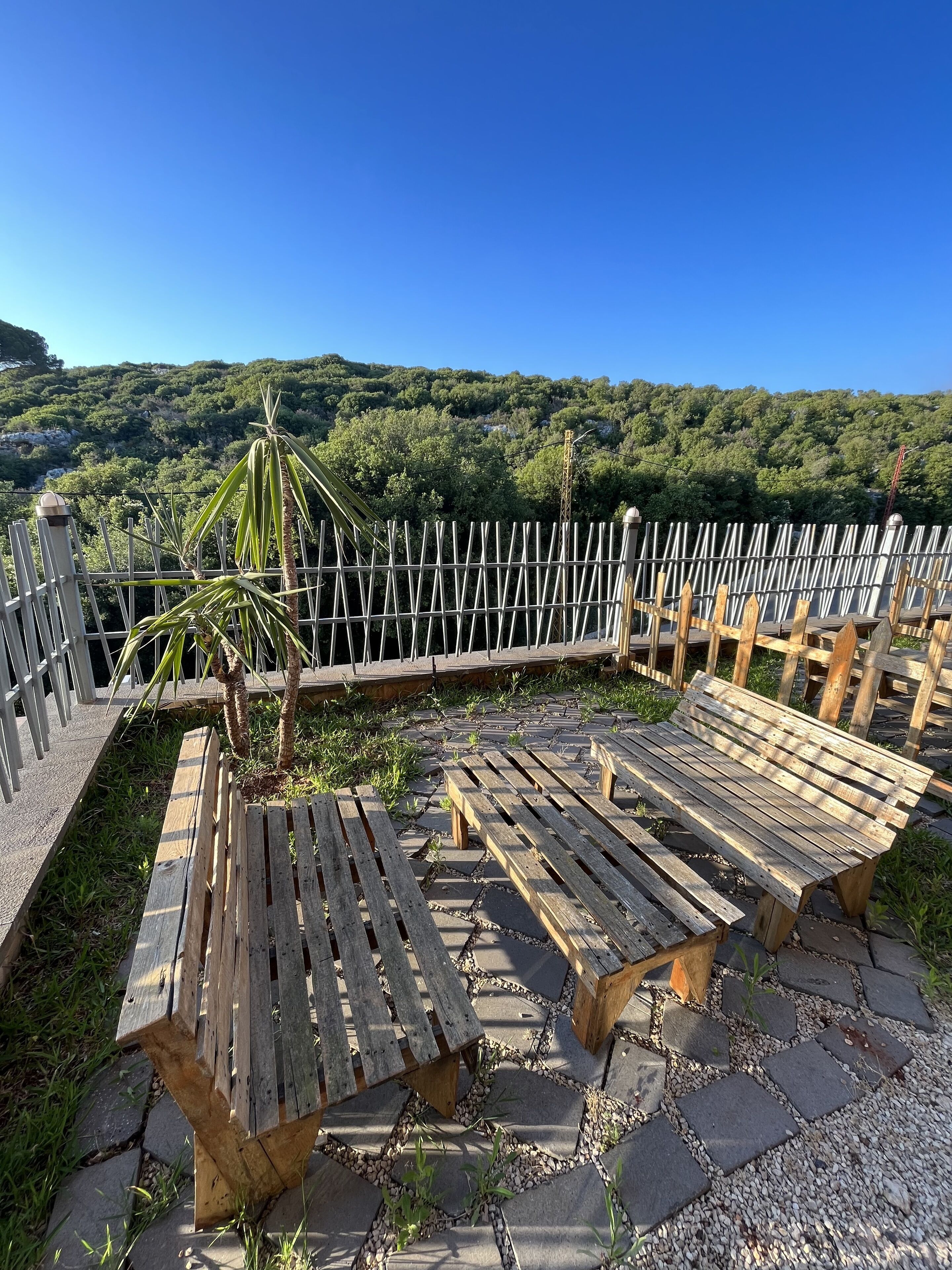 A terrace with wooden benches and a metal fence, overlooking a lush green hillside.