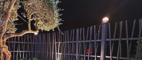 Un espacio al aire libre iluminado por la noche, con piscina, un árbol, cerca de madera y un área de descanso con faroles y cestas.