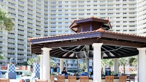 A covered outdoor seating area with tables and chairs, a pool in the background, and a tall hotel building. - Family Perfect with Lagoon pool and Crystal Sand Beach! (Panama City Beach)