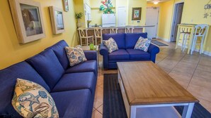 A living room with two blue sofas, a wooden coffee table, and a kitchen area in the background. - Family Perfect with Lagoon pool and Crystal Sand Beach! (Panama City Beach)