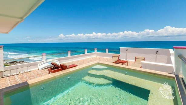 Una piscina en la azotea con agua azul cristalina, paredes blancas y sillas de descanso rojas.