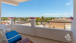 Un balcón con una silla azul acolchada, una mesa de madera y una vista despejada de edificios y palmeras a lo lejos.