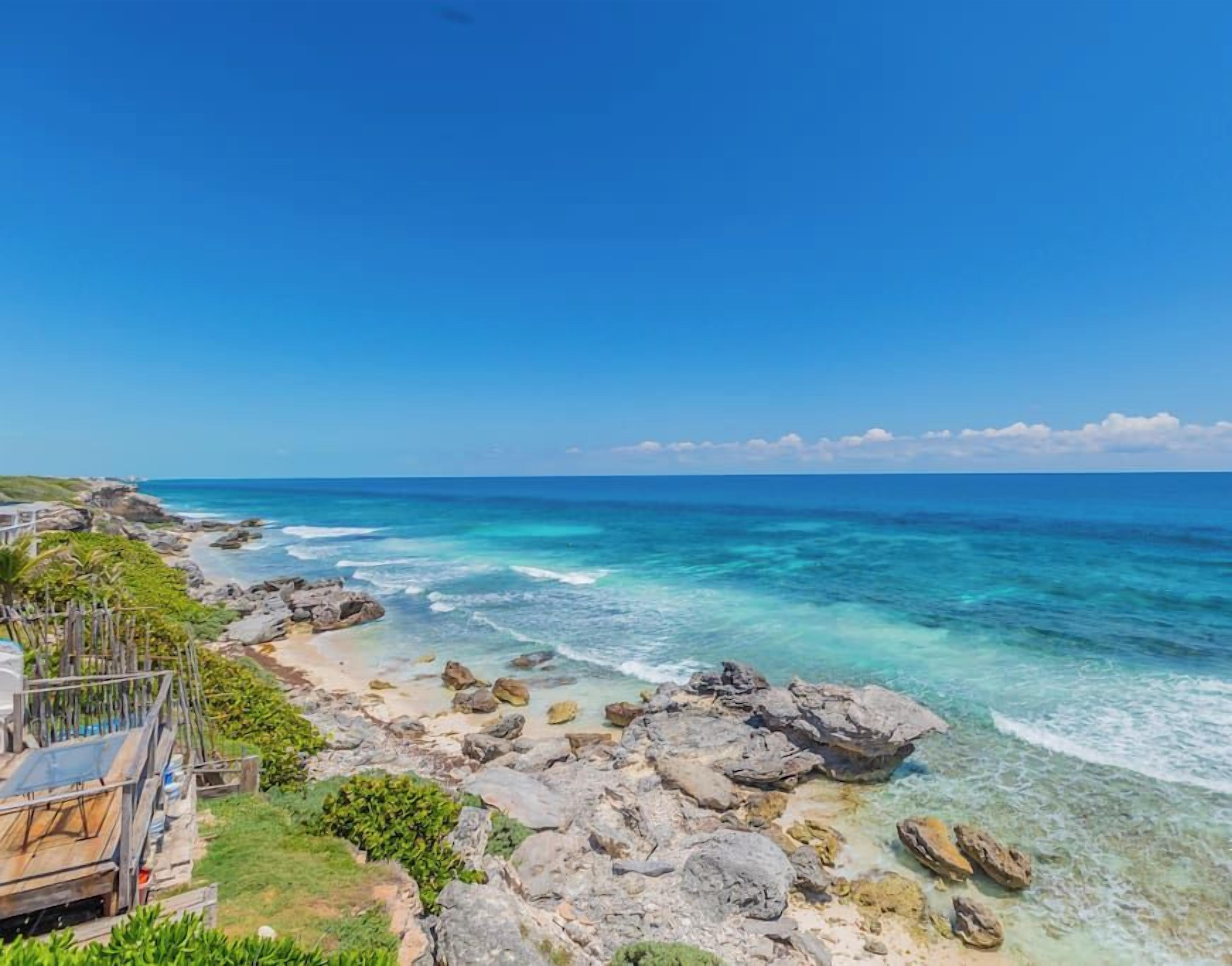A coastal view with a rocky shoreline, clear blue sea, and a wooden deck overlooking the ocean.