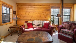 Un salón acogedor con pared de madera, un sofá rojo, un reposapiés estampado y una mesa de centro de piedra.
