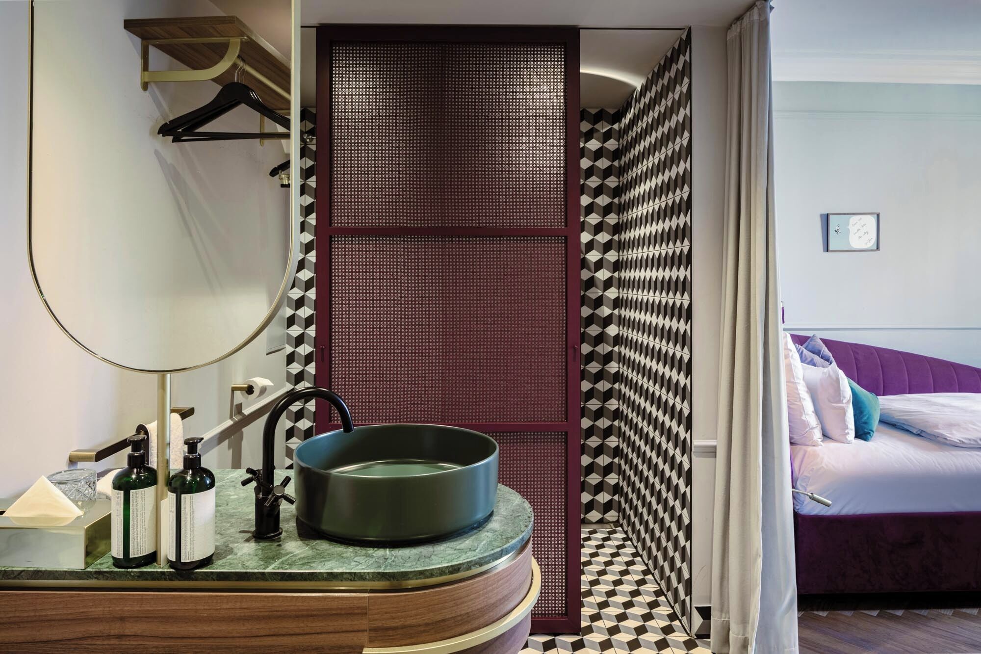 Een badkamer met een ronde spiegel, een wastafel en een toilet. In de aangrenzende kamer staat een bed met een paars hoofdeinde en kussens.