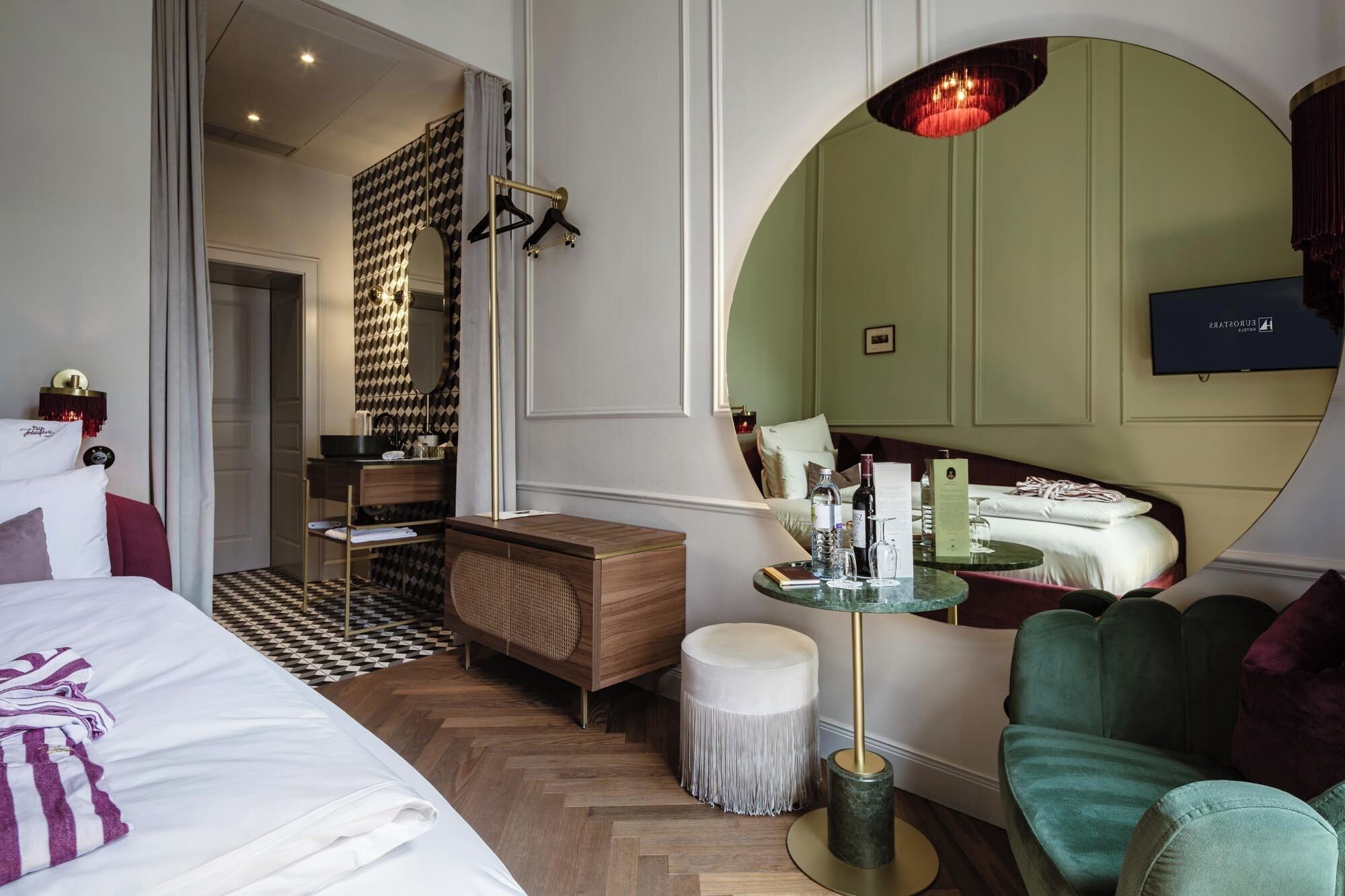 Ein Hotelzimmer mit einem Bett, einem runden Spiegel, einem Waschtisch und einem runden Tisch mit einer Glasplatte sowie einem grünen Sessel.