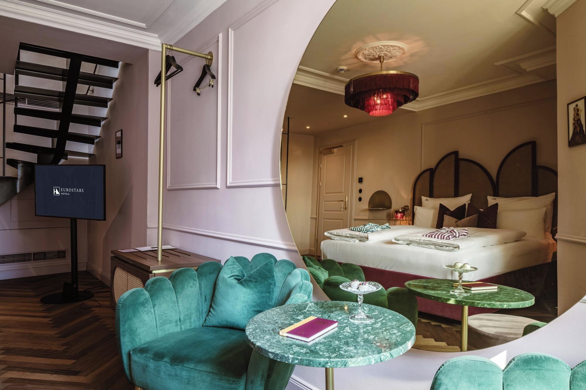 Ein Hotelzimmer mit Bett, Fernseher, einem grünen Sessel und einem runden Tisch mit Buch und Glas.