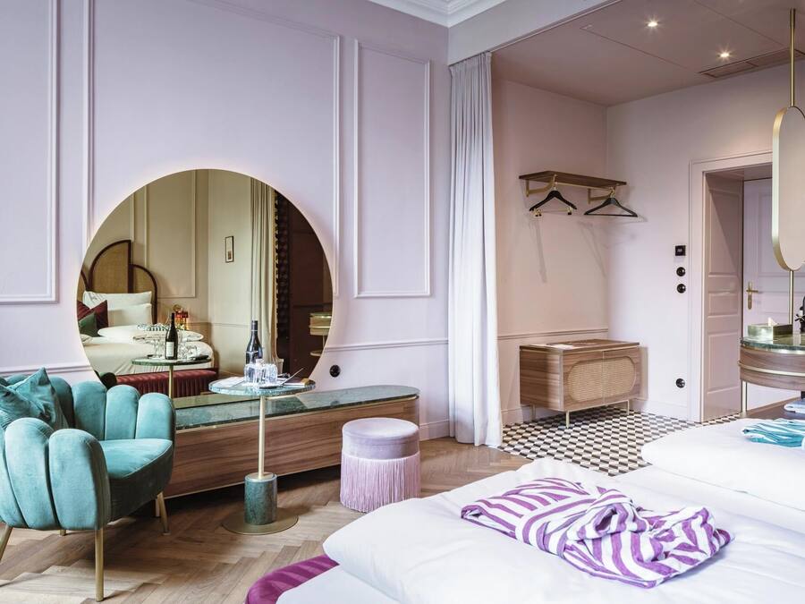 A modern bedroom with a large bed, a round mirror, a vanity with a sink, and a seating area with a chair and ottoman.