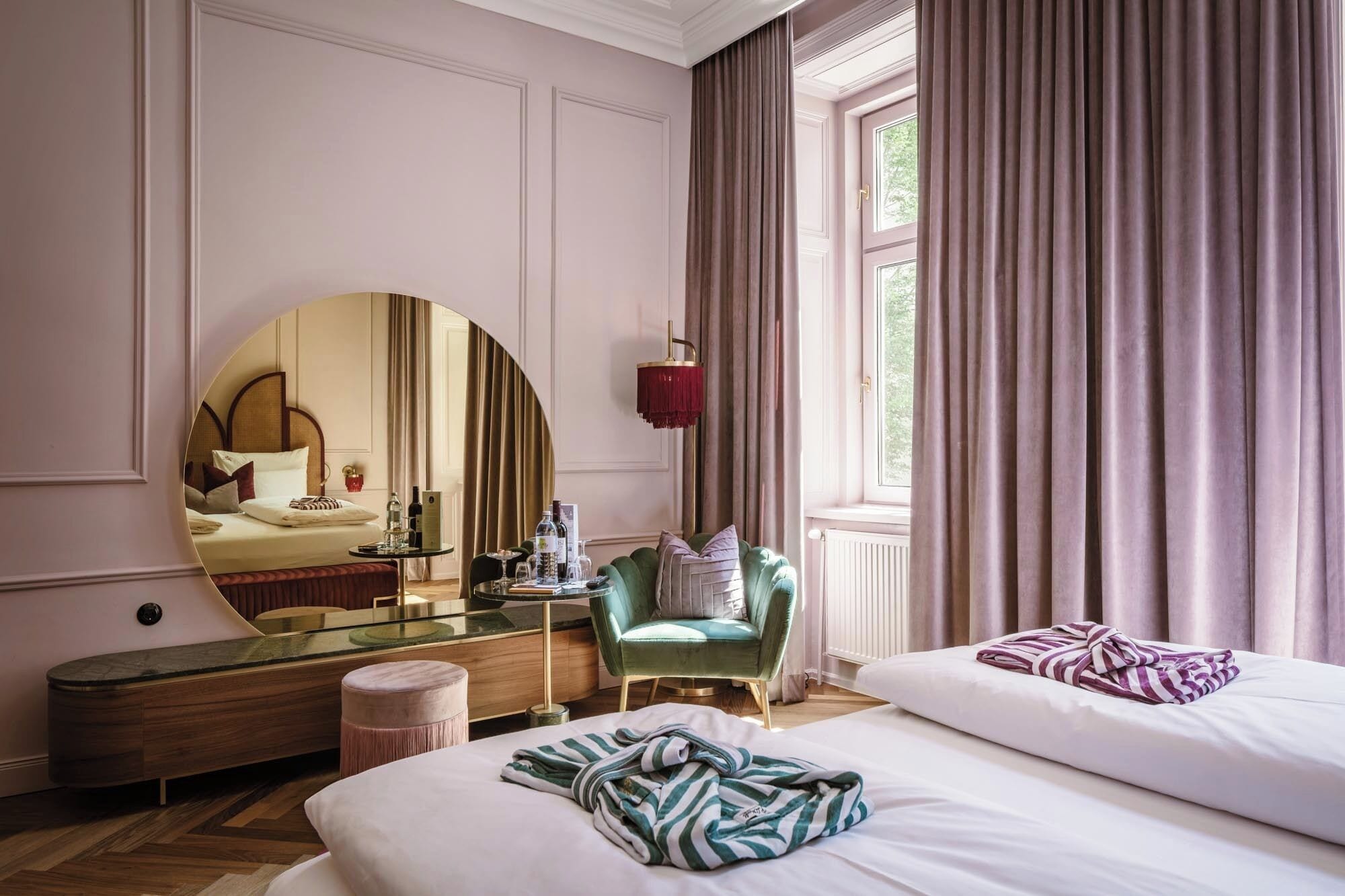 A hotel room with a large round mirror, a green armchair, a bed with a striped blanket, and a window with curtains.