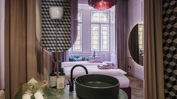 Habitación de hotel con cama, un espejo redondo, lavamanos con grifo y ventana con vitrales.