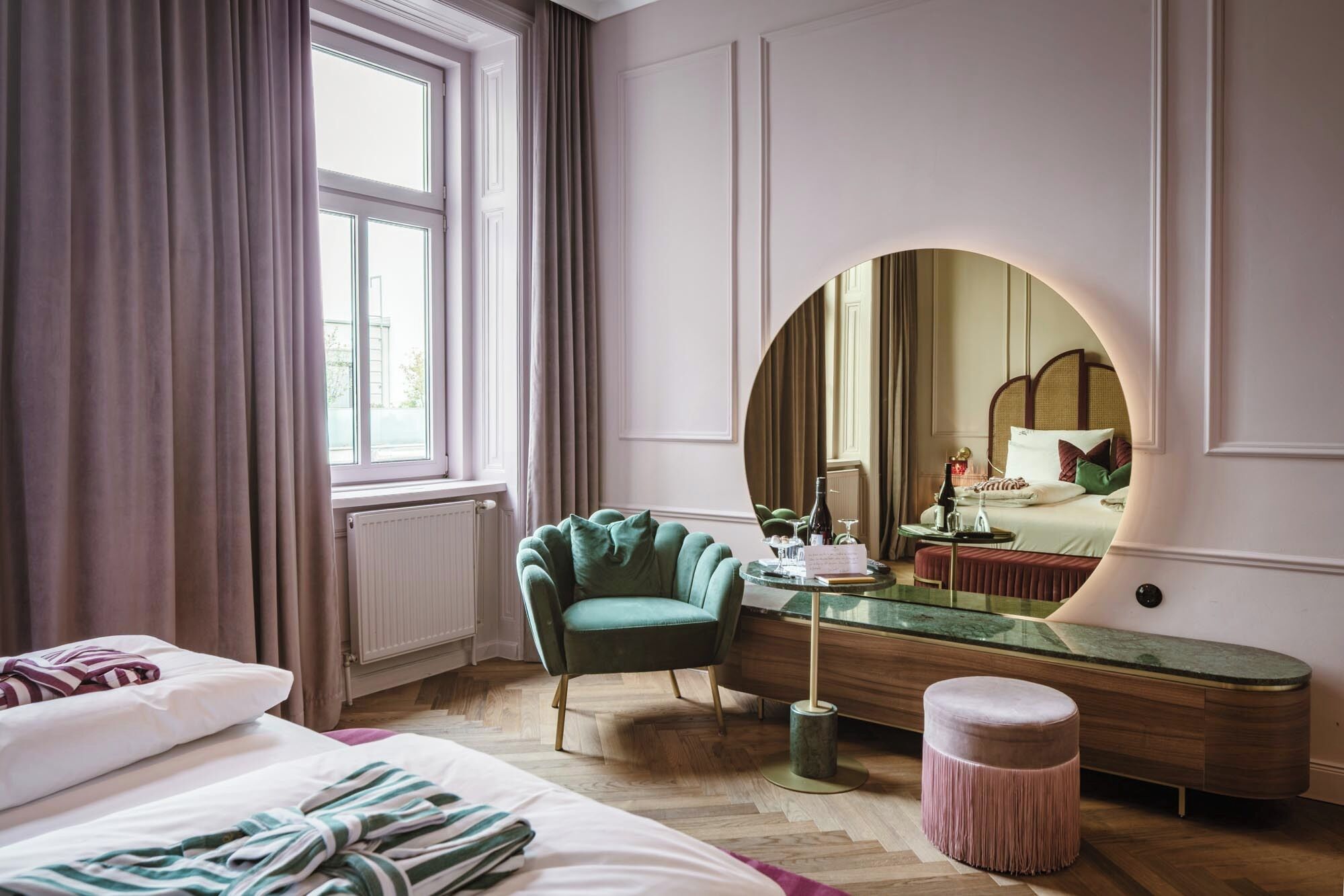 Habitación de hotel con un ventanal grande, un sillón verde, un espejo redondo, una mesita de noche y una cama con manta a rayas.