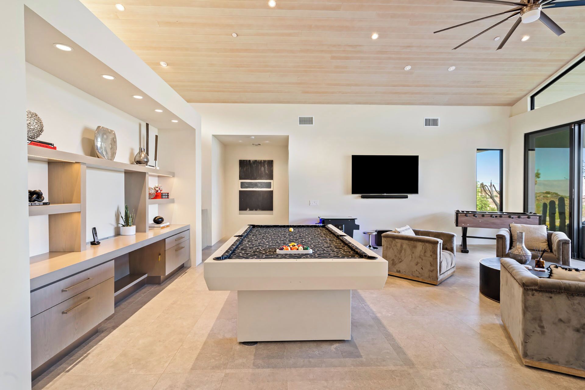 A modern living room with a pool table, a flat-screen TV, and a large window offering a view of the outdoors.