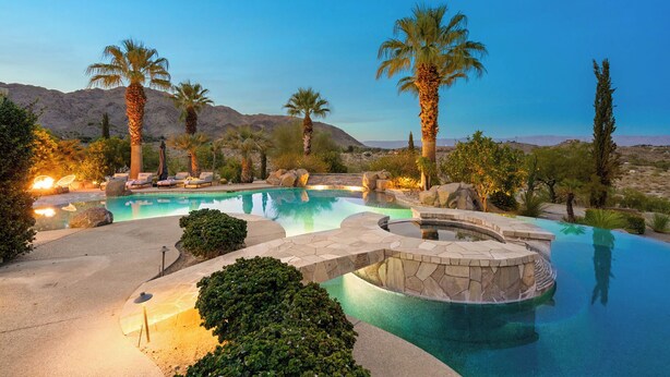 A swimming pool with a stone deck, surrounded by palm trees and rocks, set against a mountainous backdrop.