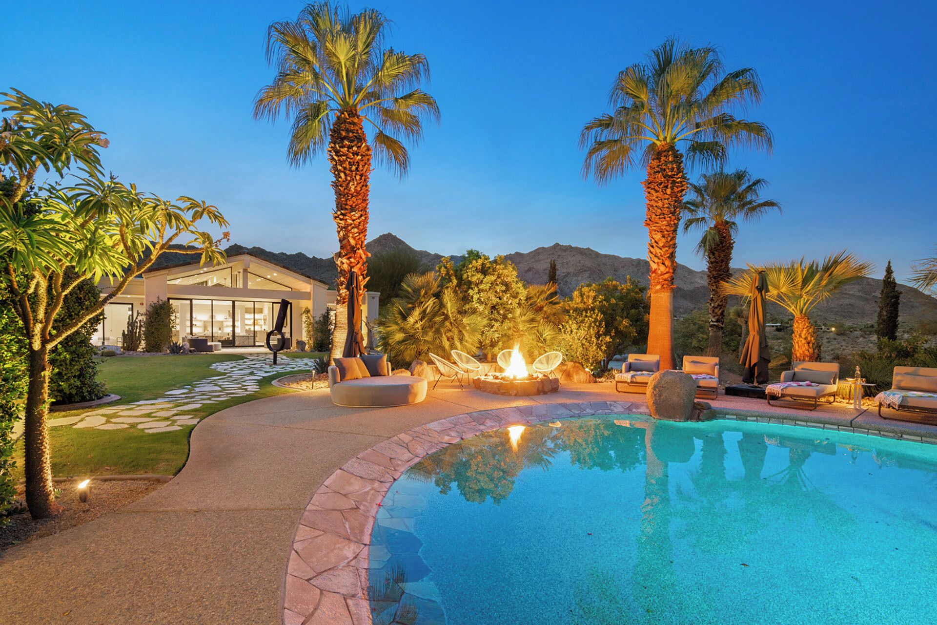 A swimming pool with lounge chairs and a fire pit, surrounded by palm trees and a mountain backdrop.