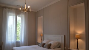 A bedroom with a bed, two bedside lamps, a chandelier, and a window with sheer curtains. - La Maison d'Antoine (Rivesaltes)