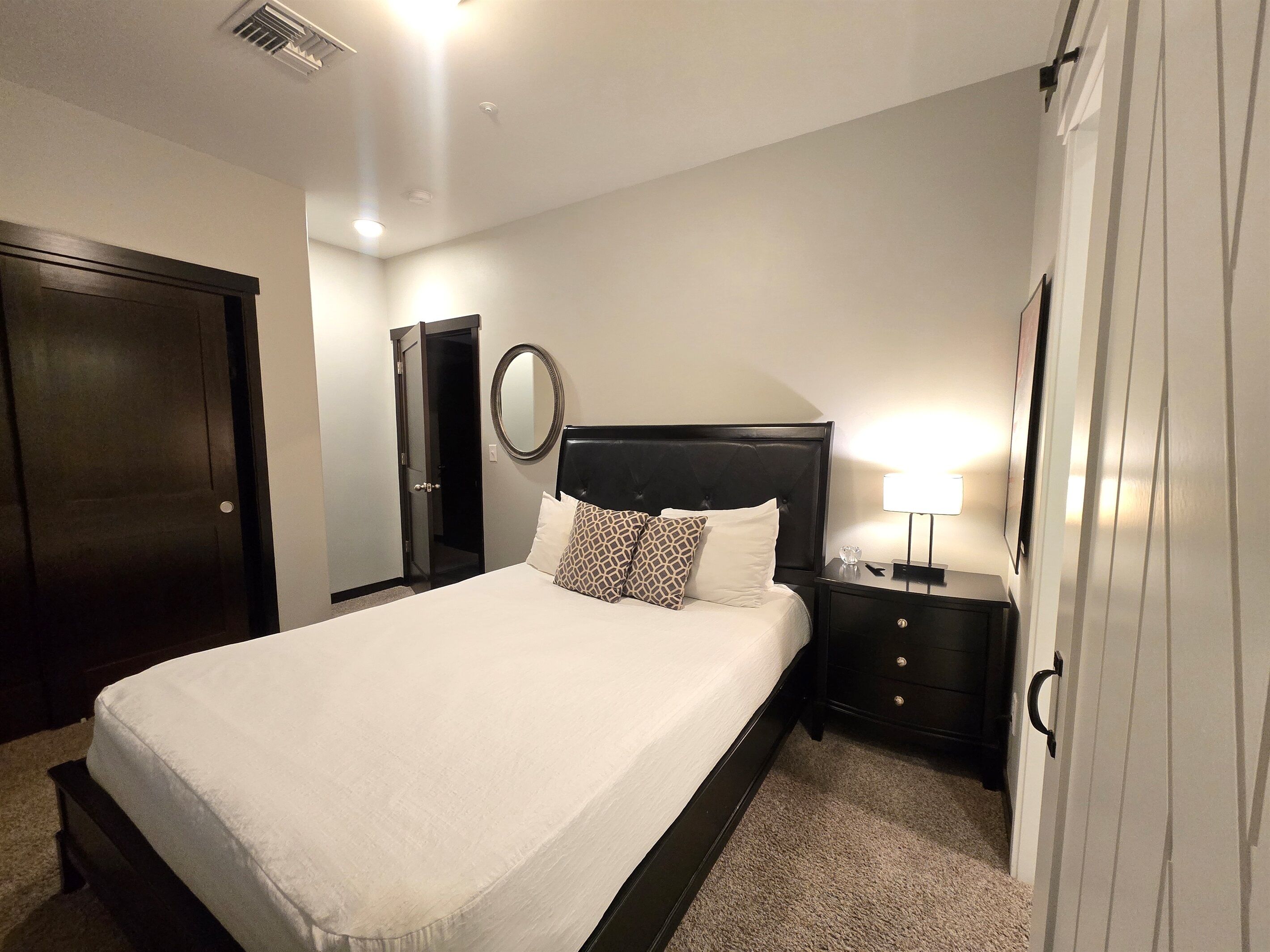 A neatly arranged bedroom with a large bed, a bedside table with a lamp, a mirror, and a door.