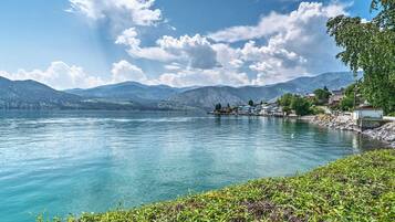 Blick auf ein Seeufer mit klarem Wasser, Bergen im Hintergrund und einer kleinen Anlegestelle mit Gebäuden.