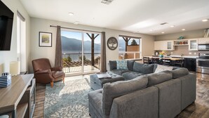A spacious living area with a large gray sectional sofa, a brown leather chair, and a wooden dining table. A kitchen with white cabinets and stainless steel appliances is visible in the background. - Majestic Views / Lake Chelan / Pool and Spas! (Manson)