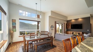 Un ampio soggiorno con un grande tavolo da pranzo, sedie in legno e un divano. C'è una TV montata a parete e una finestra con vista sull'esterno.