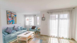 A living room with a blue sofa, a white coffee table, and a colorful rug. There is a painting on the wall and a window with sheer curtains. - Suncastle Lovely Attic! (Alicante/Alacant)