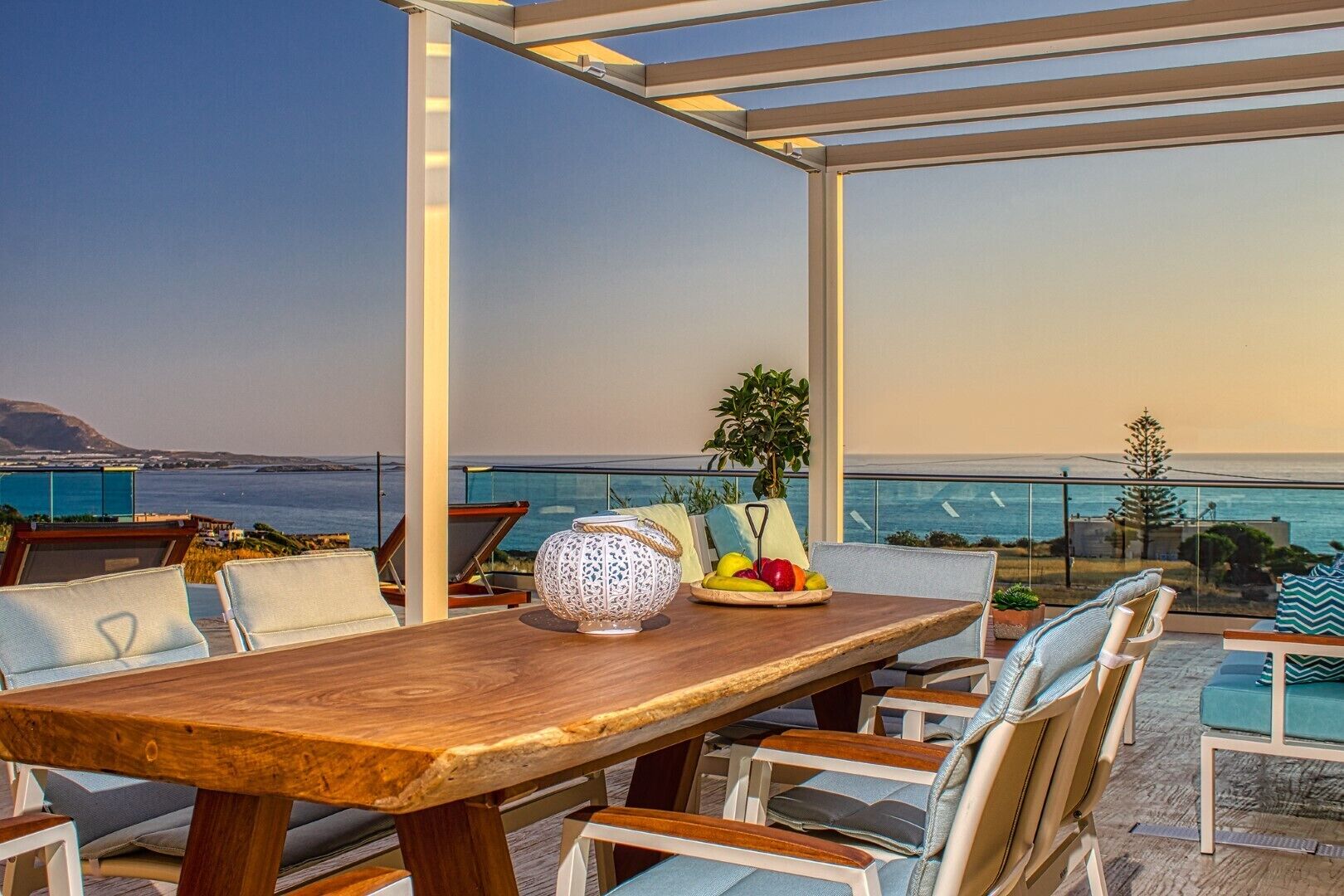 Une table à manger en bois, avec des chaises, un saladier décoratif et une assiette de fruits, sur un balcon offrant une vue sur la mer.