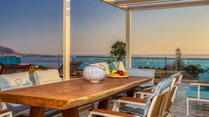 Une table à manger en bois, avec des chaises, un saladier décoratif et une assiette de fruits, sur un balcon offrant une vue sur la mer.