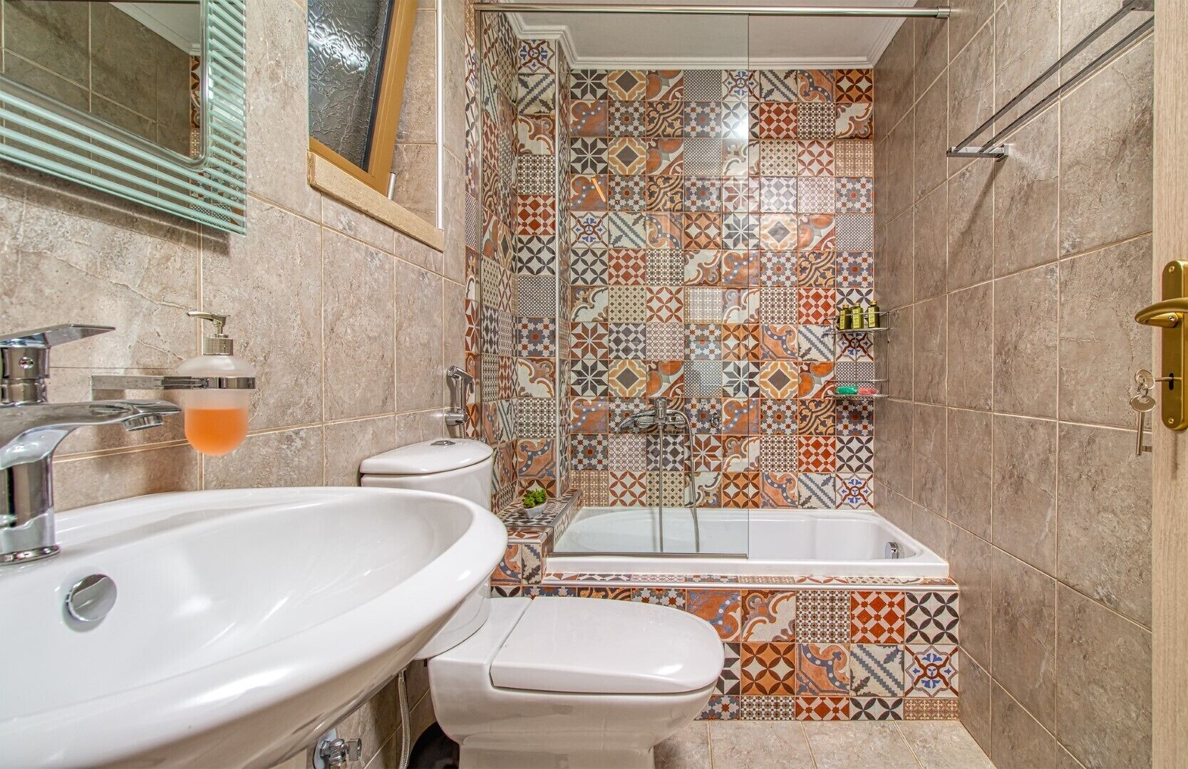 Un baño con lavamanos, inodoro y bañera. Las paredes están cubiertas de azulejos con un diseño decorativo.
