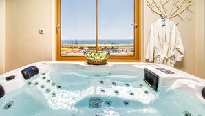 Un bain à remous avec vue sur la plage, visible à travers une grande fenêtre, un panier rempli de produits de toilette et un porte-serviettes avec un peignoir.