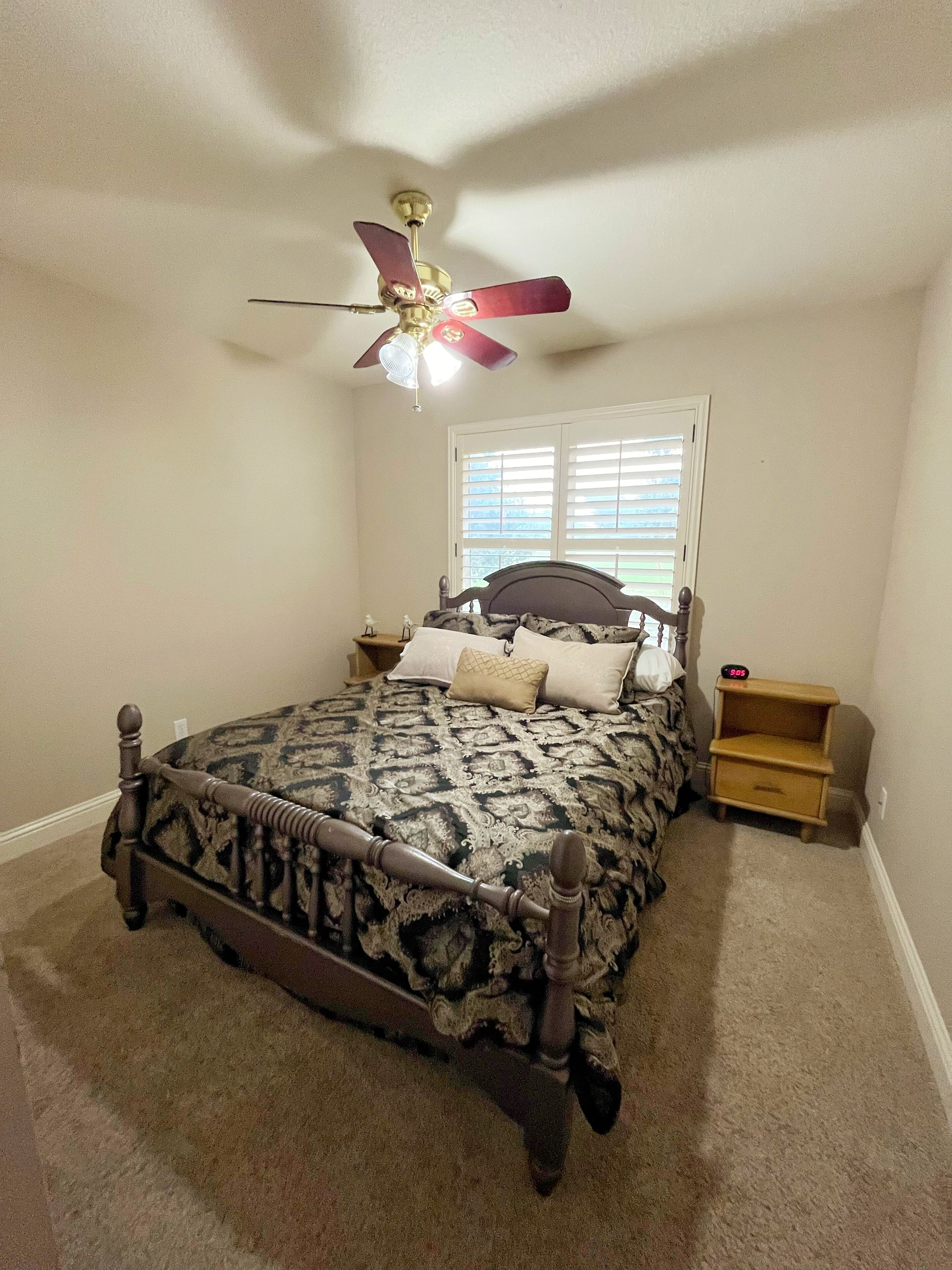 A bedroom with a bed, a nightstand, a wooden dresser, a ceiling fan, and a window with blinds.