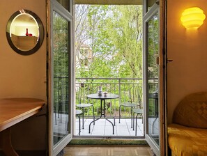 An open door leading to a balcony with a table and chairs, a round mirror on the wall, and a bed with patterned bedding.
