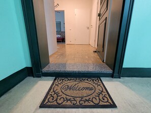 A hallway with a welcome mat leading to a bedroom with a bed and a dresser.