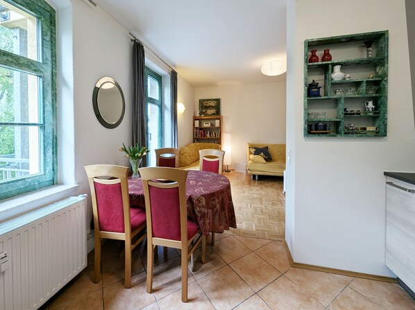 A dining area with a table and chairs, a radiator, a window with curtains, a bookshelf, and a green shelf with various items.