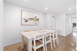 Een eetgedeelte met een houten tafel en stoelen, een ingelijst schilderij aan de muur en een witte deur.