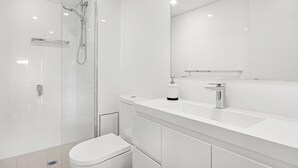 Une salle de bain moderne avec une douche à parois de verre, des toilettes blanches et un grand miroir au-dessus d’un lavabo blanc.