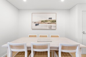A dining area with a wooden table, white chairs, and a framed landscape painting above.