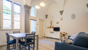A dining area with a table and chairs, a sofa, a TV on a cabinet, and a window with a view of greenery. - Le Volte by Mmega (Florence)