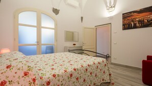 A bedroom with a floral bedspread, a large arched window, a red chair, and a cityscape painting on the wall. - Le Volte by Mmega (Florence)