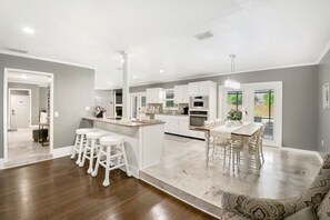 A modern kitchen with white cabinetry, a central island with bar stools, and a dining area with a table and chairs. - Lakeland Highlands Oasis - 3BR Heated Pool Home (Lakeland)