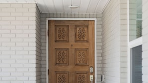 A wooden door with intricate carvings, a white brick wall, and a checkered floor leading to a porch with a gray sofa and a blue and white patterned pillow.